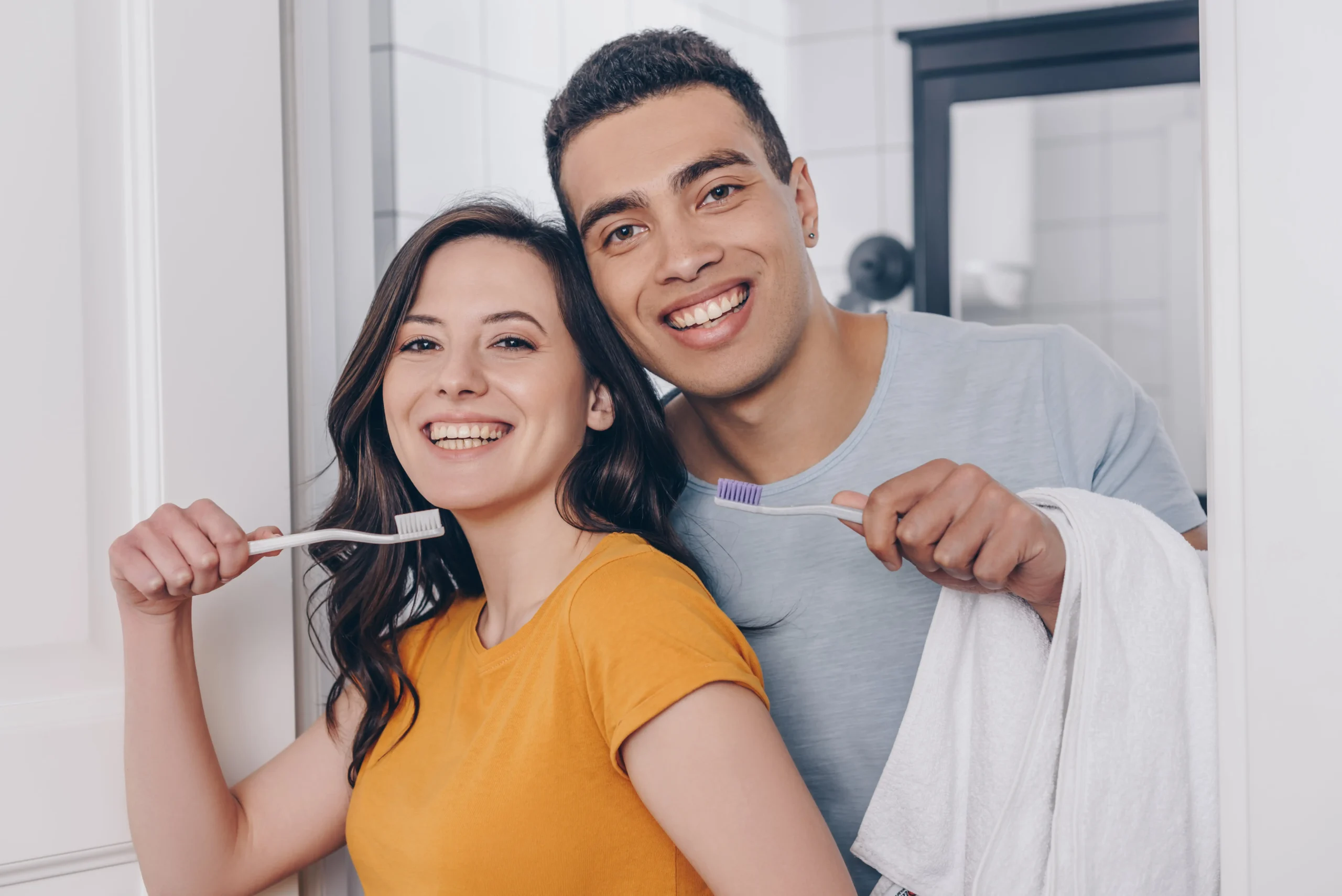 A man and a woman starting at the mirror smiling while holding their toothbrushes.