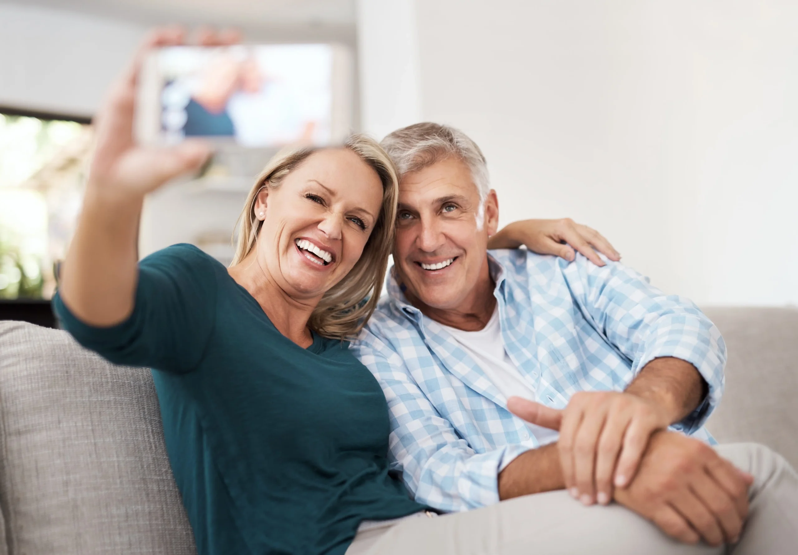 A woman and man with perfect teeth seated on a couch trying to take a photo with their phone.